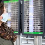 a woman wearing a face mask is standing in front of digital boards with all flight information at the airport