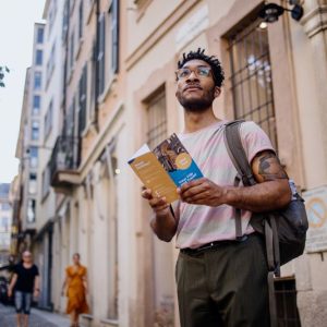 A photo of a male tourist, standing in the street of a city in Europe, holding a brochure.